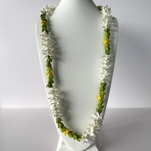 May include: A white lei necklace with green and yellow accents, displayed on a white mannequin. The necklace features white flower-like elements interspersed with green and yellow foliage, creating a vibrant and natural aesthetic.