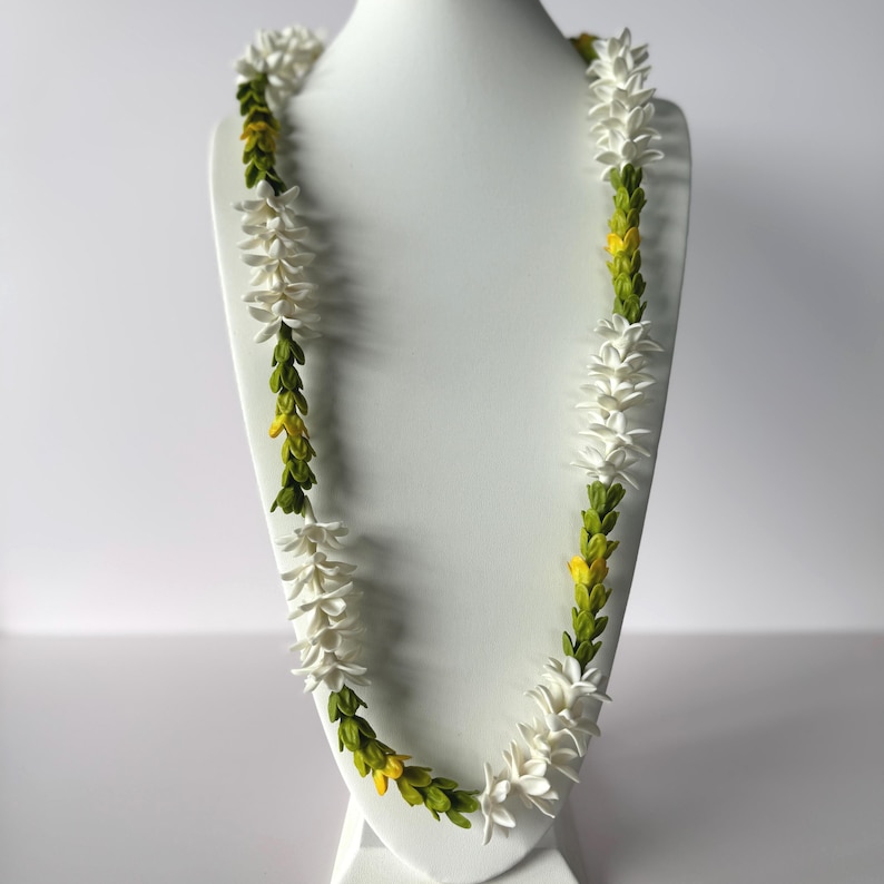 May include: A white lei necklace with green and yellow accents, displayed on a white mannequin. The necklace features white flower-like elements interspersed with green and yellow foliage, creating a vibrant and natural aesthetic.