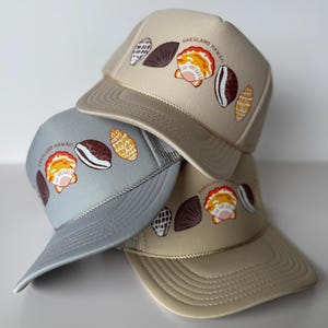 May include: Three trucker hats stacked, each in a different neutral color: tan, gray, and beige. Each hat features a seashell design and the text "HAKULAND HAWAII" in a semi-circle above the seashells. The hats have a mesh back and a curved brim.