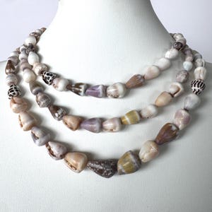 May include: Three strands of seashell necklaces displayed on a white mannequin. The shells are in shades of white, tan, brown, and purple. The shells are strung together to create a layered necklace.