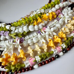 May include: A collection of colorful leis, featuring various floral designs in white, yellow, pink, and purple. Some leis incorporate wooden beads and pearls, creating a vibrant and decorative display.