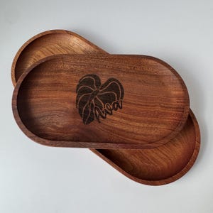 May include: Two wooden oval trays with a dark brown finish. The top tray has a leaf design and the word "Ava" etched into the wood. The trays are stacked, showcasing their shape and the wood grain.