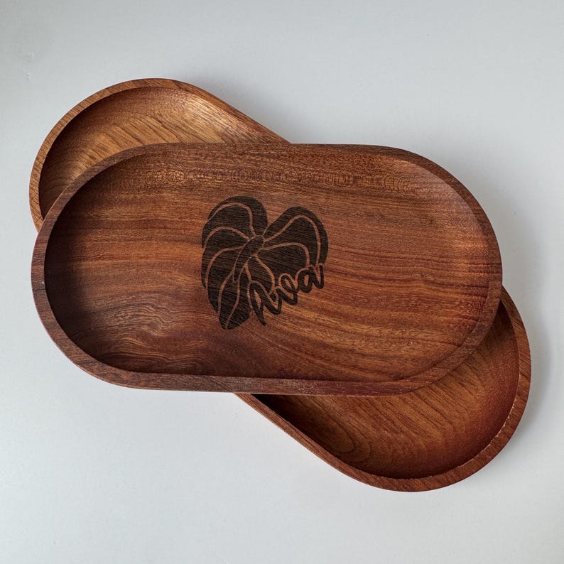 May include: Two wooden oval trays with a dark brown finish. The top tray has a leaf design and the word "Ava" etched into the wood. The trays are stacked, showcasing their shape and the wood grain.