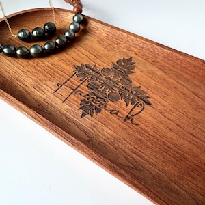 Może przedstawiać: Drewniana taca w kolorze czerwonobrązowym z wygrawerowanym imieniem "Hannah" kursywą i kwiatowym wzorem. Złoty naszyjnik z ciemnoszarymi perłami i naszyjnik z koralików spoczywają na tacy. Taca jest wykonana z drewna.