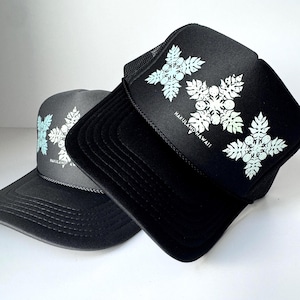 May include: Two black trucker hats with a floral design in light blue and white. The hats have the text "HAKULAND HAWAII" printed on the front. One hat is black with a black mesh back, and the other is dark gray.