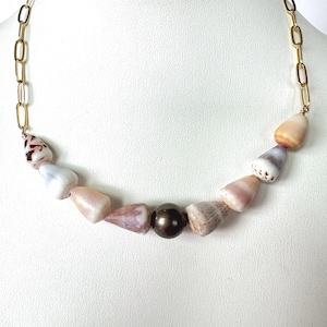 May include: A necklace featuring a variety of seashells in shades of white, pink, and brown, strung together with a gold chain. A dark brown pearl is centered in the design. The necklace is displayed against a white background.