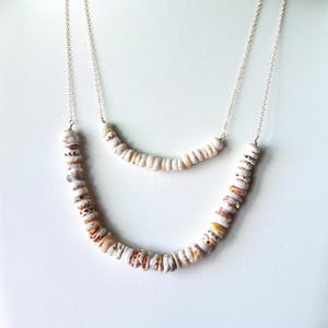 May include: Two layered necklaces with white and tan shell beads. The necklaces have a delicate silver chain. The beads have brown and orange spots. The necklaces are displayed on a white background.
