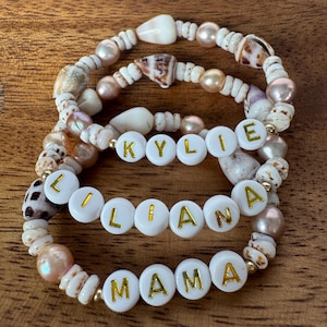 May include: Three handmade bracelets with white, gold, and shell beads. One bracelet spells out "KYLIE", another "LIANA", and the third "MAMA" in gold letters on white beads. The bracelets are arranged on a wooden surface.