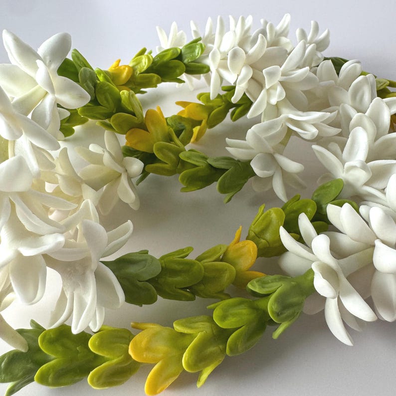May include: Close-up of a white flower lei with green and yellow leaves. The lei is made of delicate, white flowers and vibrant green and yellow leaves, creating a natural and elegant design. The floral arrangement is perfect for a special occasion.