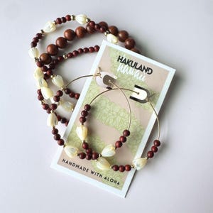 Könnte beinhalten: Goldene Creolen und Halsketten mit tropischem Thema. Die Ohrringe und Halsketten verfügen über dunkelbraune Holzperlen, weiße muschelförmige Perlen und goldfarbene Akzente. Eine Karte mit dem Text "HAKULAND Hawaii" und "HANDMADE WITH ALOHA" ist ebenfalls vorhanden.