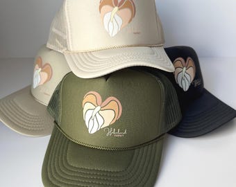 May include: A collection of trucker hats in various colors: beige, tan, olive green, and navy blue. Each hat features a heart-shaped graphic with a floral design in shades of peach, brown, and white, along with the text "HokuLand Hawaii".