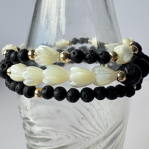 May include: Two stacked bracelets with black lava beads, cream-coloured flower-shaped beads, and gold-coloured accent beads. The bracelets are displayed on a clear glass surface.