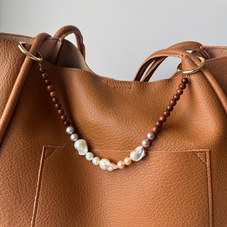 May include: A tan leather tote bag with a decorative beaded strap. The strap features wooden beads and irregularly shaped pearls in white, pink, and peach tones. Gold-colored rings attach the strap to the bag.