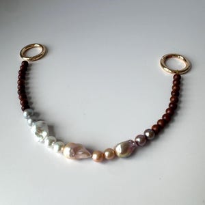 May include: A beaded bag strap featuring a mix of baroque pearls and dark brown wooden beads. The pearls vary in size and color, ranging from white to pink and lavender. Gold-colored metal rings are at each end.