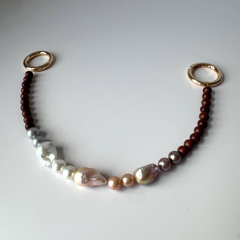 May include: A beaded bag strap featuring a mix of baroque pearls and dark brown wooden beads. The pearls vary in size and color, ranging from white to pink and lavender. Gold-colored metal rings are at each end.