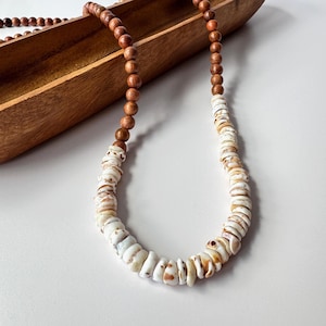 May include: A handcrafted necklace featuring alternating sections of brown wooden beads and white shell beads. The shell beads have natural brown markings. The necklace is displayed on a white surface with a wooden tray in the background.