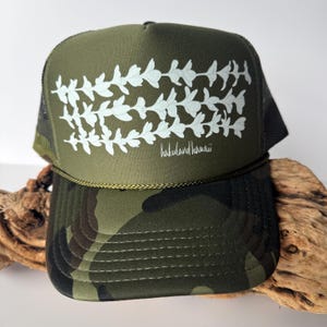 May include: An olive green trucker hat featuring a white floral lei graphic across the front panel and the text "hakuland hawaii" in a script font. The brim displays a camouflage pattern, and a thin gold cord runs along the base of the crown.