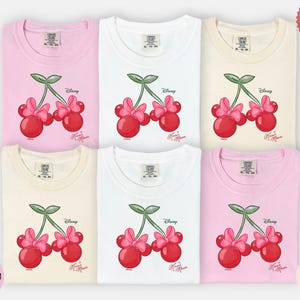 Peut inclure: Six t-shirts de couleurs vari&eacute;es : rose, blanc et ivoire. Chaque t-shirt pr&eacute;sente un motif de deux cerises rouges avec des n&oelig;uds roses et des tiges vertes, avec les logos Disney et Minnie Mouse.