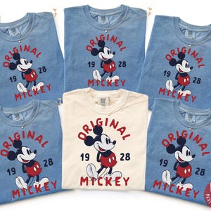 May include: A collection of blue and cream-colored t-shirts featuring a vintage-style Mickey Mouse graphic. The design includes the words "ORIGINAL," "MICKEY," and the numbers "19" and "28." Each shirt has a small silver necklace.