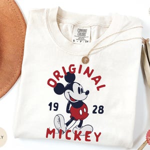 May include: Ivory-colored t-shirt featuring a graphic of Mickey Mouse with the text "ORIGINAL MICKEY 1928" in red and navy blue. The shirt is adorned with a gold necklace. The shirt is made of a soft material.