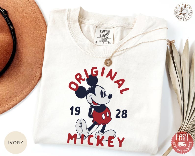 May include: Ivory-colored t-shirt featuring a graphic of Mickey Mouse with the text "ORIGINAL MICKEY 1928" in red and navy blue. The shirt is adorned with a gold necklace. The shirt is made of a soft material.