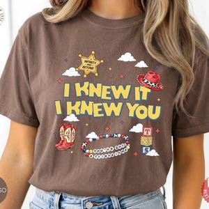 May include: A brown short-sleeve t-shirt featuring a graphic print with the text I KNEW IT I KNEW YOU, a yellow sheriff star badge, a red cowboy hat, a pair of red cowboy boots, a beaded bracelet with the text YOU'VE GOT A FRIEND IN ME, and blocks.