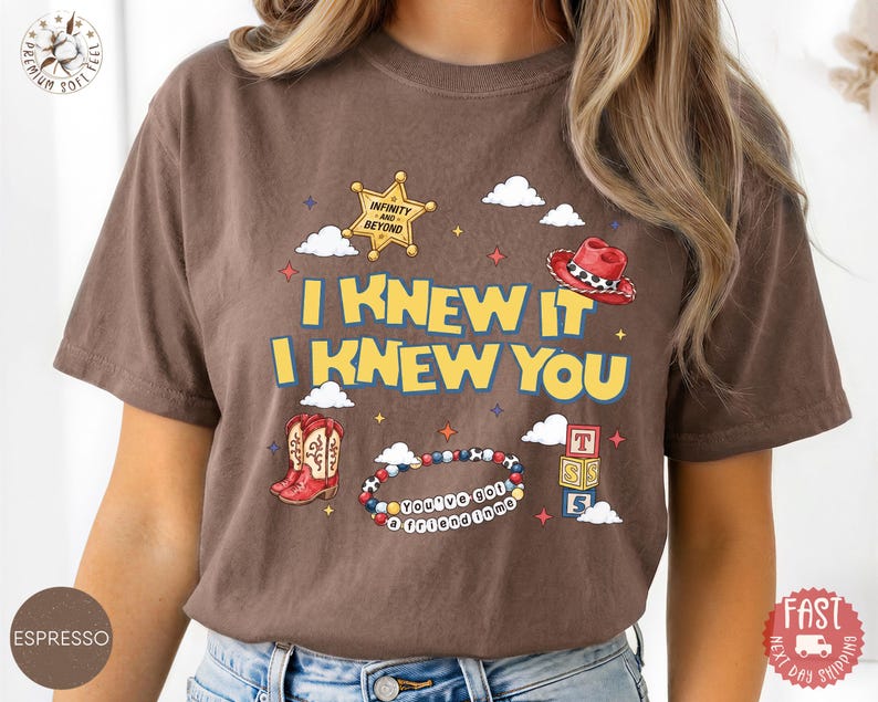 May include: A brown short-sleeve t-shirt featuring a graphic print with the text I KNEW IT I KNEW YOU, a yellow sheriff star badge, a red cowboy hat, a pair of red cowboy boots, a beaded bracelet with the text YOU'VE GOT A FRIEND IN ME, and blocks.
