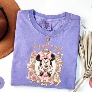 May include: A violet short-sleeved t-shirt featuring a graphic of Minnie Mouse inside an ornate gold frame with pink roses and the text I'm just a girl. The shirt has a crew neckline and a Comfort Colors label on the inside of the collar.