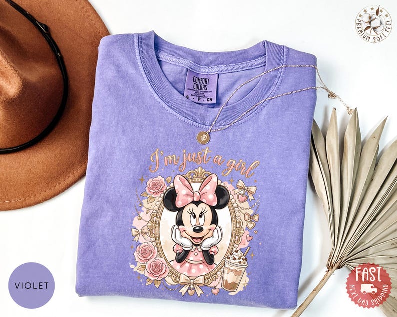 May include: A violet short-sleeved t-shirt featuring a graphic of Minnie Mouse inside an ornate gold frame with pink roses and the text I'm just a girl. The shirt has a crew neckline and a Comfort Colors label on the inside of the collar.