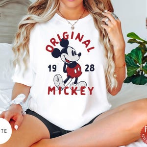 May include: White t-shirt featuring a classic Mickey Mouse design. The graphic includes the words "ORIGINAL MICKEY" in red, with "19 28" below the character. The shirt is a crew neck style.