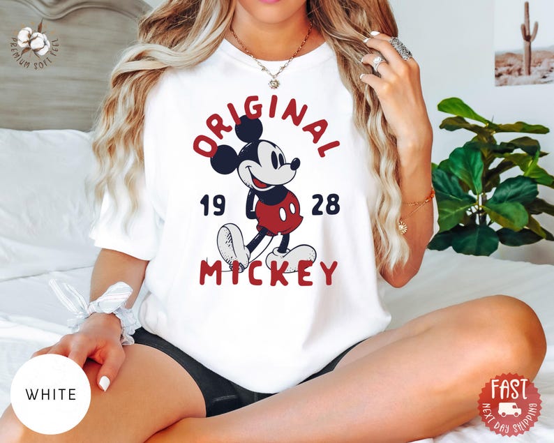 May include: White t-shirt featuring a classic Mickey Mouse design. The graphic includes the words "ORIGINAL MICKEY" in red, with "19 28" below the character. The shirt is a crew neck style.