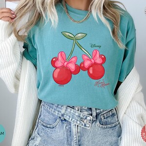 Peut inclure: T-shirt vert d'eau avec un motif de cerises de dessin anim&eacute;, des n&oelig;uds roses et le texte "Disney Minnie Mouse". Le t-shirt est fait d'une mati&egrave;re douce.