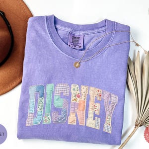 May include: A violet short-sleeved t-shirt with a crew neckline featuring the word DISNEY in a patchwork-style applique. The letters are composed of various pastel-colored fabrics with floral, plaid, and solid patterns. A thin gold chain necklace rests on the shirt.