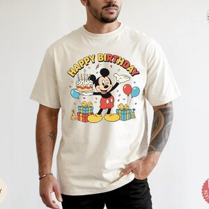 K&ouml;nnte beinhalten: Cremefarbenes T-Shirt mit einer Mickey Mouse-Grafik, die eine Geburtstagstorte, Luftballons und Geschenke h&auml;lt. Der Text "HAPPY BIRTHDAY" ist oben aufgebogen. Das Design ist in Rot, Gelb, Schwarz und Blau gehalten.