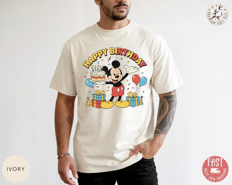 K&ouml;nnte beinhalten: Cremefarbenes T-Shirt mit einer Mickey Mouse-Grafik, die eine Geburtstagstorte, Luftballons und Geschenke h&auml;lt. Der Text "HAPPY BIRTHDAY" ist oben aufgebogen. Das Design ist in Rot, Gelb, Schwarz und Blau gehalten.