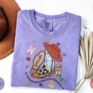 May include: A violet short-sleeve t-shirt featuring a graphic print of a cowboy boot with a cow print pattern, a red hat, the name Taylor, the number 13, and stacked blocks with the letters T, S, and S, surrounded by small floral and star icons.