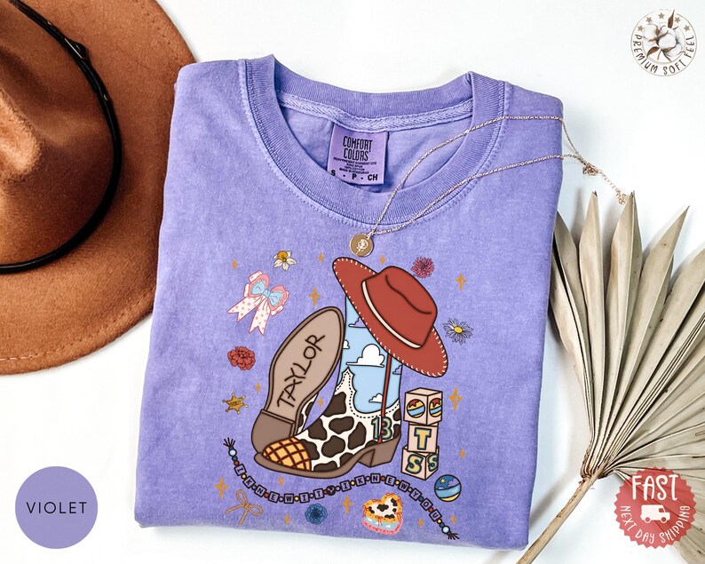May include: A violet short-sleeve t-shirt featuring a graphic print of a cowboy boot with a cow print pattern, a red hat, the name Taylor, the number 13, and stacked blocks with the letters T, S, and S, surrounded by small floral and star icons.
