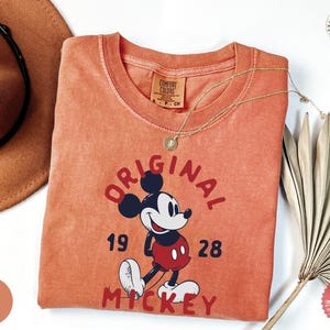 May include: A coral-colored t-shirt featuring a graphic of Mickey Mouse with the text "ORIGINAL MICKEY 1928". The shirt has a crew neck and short sleeves. A gold necklace with a pendant is draped across the shirt. A brown hat and dried plant are in the background.
