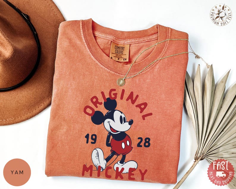 May include: A coral-colored t-shirt featuring a graphic of Mickey Mouse with the text "ORIGINAL MICKEY 1928". The shirt has a crew neck and short sleeves. A gold necklace with a pendant is draped across the shirt. A brown hat and dried plant are in the background.
