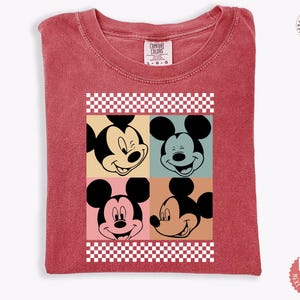 Peut inclure: T-shirt rouge avec un motif de style pop art repr&eacute;sentant le visage de Mickey Mouse en quatre expressions diff&eacute;rentes. Le motif est encadr&eacute; d'un motif &agrave; carreaux. Le t-shirt a un col rond et des manches courtes.