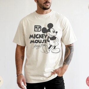 May include: Cream-coloured t-shirt with a vintage Mickey Mouse graphic. The design features Mickey Mouse walking, with the words "Mickey Mouse" in a bold font. The shirt has short sleeves and a relaxed fit.