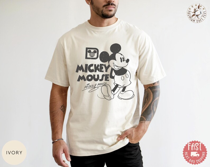 May include: Cream-coloured t-shirt with a vintage Mickey Mouse graphic. The design features Mickey Mouse walking, with the words "Mickey Mouse" in a bold font. The shirt has short sleeves and a relaxed fit.