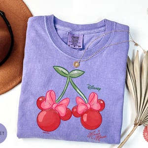 Peut inclure: Un t-shirt violet avec un motif de cerises Minnie Mouse. Le graphisme comprend des cerises rouges avec des n&oelig;uds roses, des feuilles vertes et le logo Disney. Un collier en or repose sur le t-shirt. Le t-shirt est en mati&egrave;re douce.