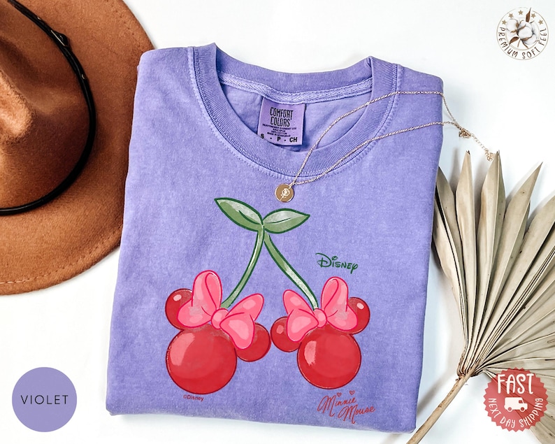 Peut inclure: Un t-shirt violet avec un motif de cerises Minnie Mouse. Le graphisme comprend des cerises rouges avec des n&oelig;uds roses, des feuilles vertes et le logo Disney. Un collier en or repose sur le t-shirt. Le t-shirt est en mati&egrave;re douce.