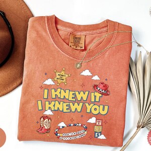 May include: A terracotta-colored t-shirt featuring a graphic print with the text I KNEW IT I KNEW YOU, a sheriff badge labeled INFINITY AND BEYOND, a red cowboy hat, a pair of western boots, a beaded bracelet, and three stacked alphabet blocks.
