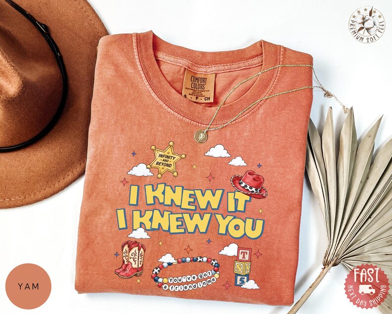 May include: A terracotta-colored t-shirt featuring a graphic print with the text I KNEW IT I KNEW YOU, a sheriff badge labeled INFINITY AND BEYOND, a red cowboy hat, a pair of western boots, a beaded bracelet, and three stacked alphabet blocks.