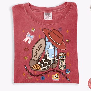 May include: A red short-sleeve t-shirt featuring a graphic print of a cow-print cowboy boot, a brown cowboy hat, alphabet blocks spelling BTS, a bow, and a beaded string. The word TAYLOR is printed on the boot, and the number 13 is visible.