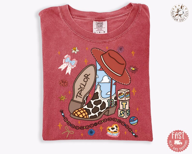 May include: A red short-sleeve t-shirt featuring a graphic print of a cow-print cowboy boot, a brown cowboy hat, alphabet blocks spelling BTS, a bow, and a beaded string. The word TAYLOR is printed on the boot, and the number 13 is visible.