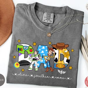 May include: A charcoal gray t-shirt featuring the word DAD in large letters decorated with Toy Story character patterns. Buzz Lightyear, Woody, and small stars are printed on the front. The names Olivia, Jonathan, and Emma are written below.