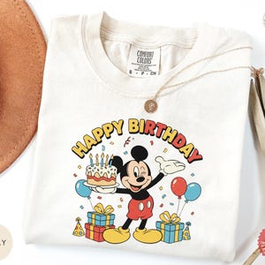 K&ouml;nnte beinhalten: Elfenbeinfarbenes T-Shirt mit einem farbenfrohen Mickey Mouse-Geburtstagsmotiv. Das Design zeigt einen Kuchen, Luftballons und den Schriftzug "HAPPY BIRTHDAY". Das T-Shirt hat einen Rundhalsausschnitt und kurze &Auml;rmel.